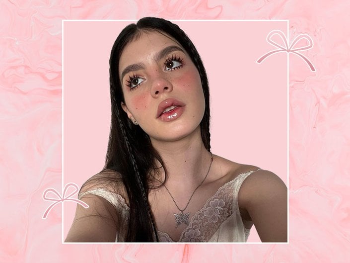Selfie of a person with long dark hair and coquette style makeup looking up to the side of the camera, collaged onto a light pink background