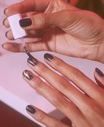 Photo of a person’s hands. Their fingernails are painted  black, and they are applying a top coat of glittery nail polish