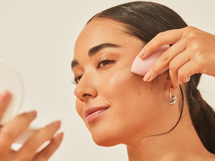 Photo of a young person using a makeup blending sponge to apply foundation while looking into a compact mirror.