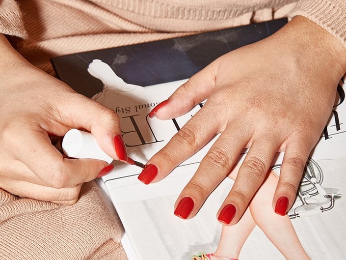 Close-up of someone painting their nails with red nail polish while their hand rests on a magazine