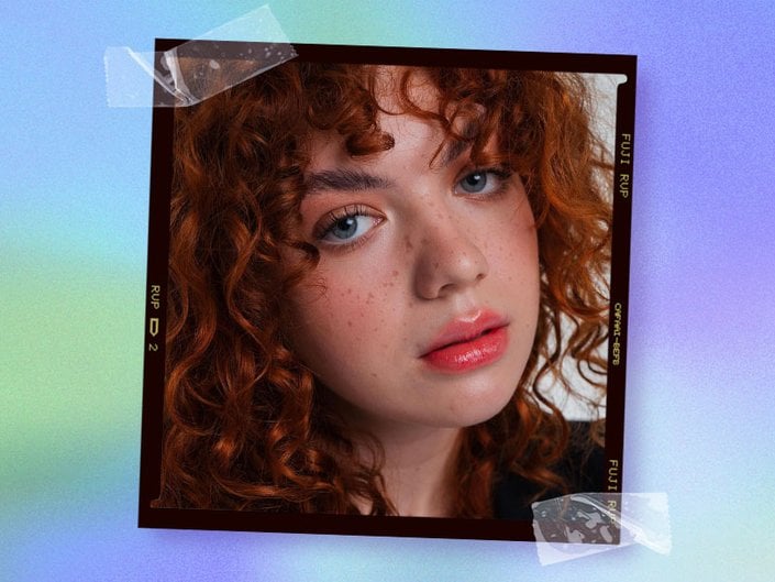 Close-up image of a person with curly red hair, blue eyes, and fake freckles looking at the camera, collaged onto a green and purple tie-dye background.
