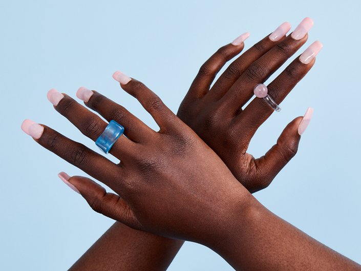 Picture of two hands with long, light pink acrylic nails and colorful rings against a blue background