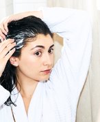 Person with dark hair wearing a white robe. Their hair is wet and they are rubbing product into it with both hands.