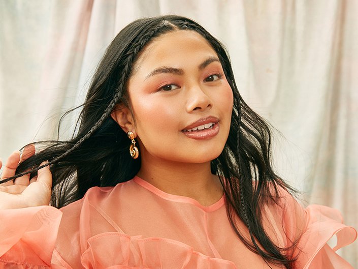 person with long dark hair with two face-framing braids and peach-colored makeup matching a peach-colored shirt